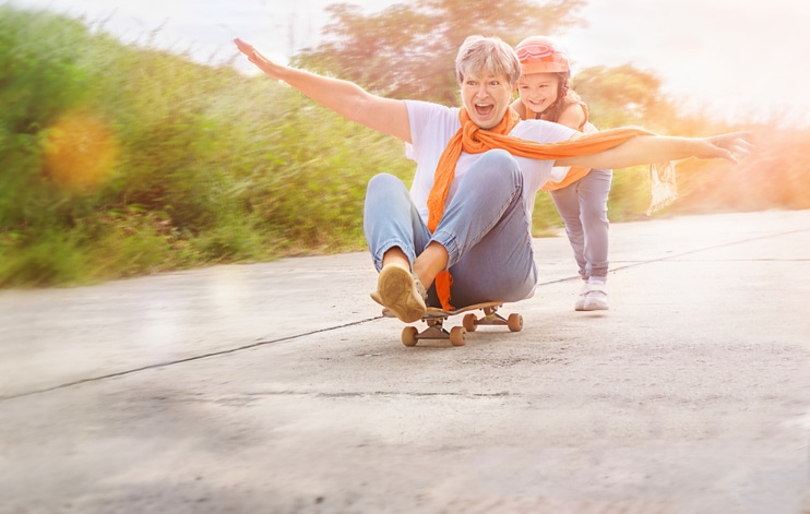 An older woman with gray hair, wearing an orange scarf and light blue jeans, sits joyfully on a skateboard, her arms spread wide. A young child with a helmet and a big smile stands behind her, pushing the skateboard. The scene takes place outdoors on a sunny day, with a blurred green background and a warm, happy atmosphere.