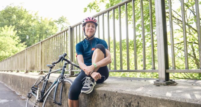 A middle-aged woman, dressed in cycling gear and a helmet, takes a break during a bike ride, sitting on a concrete ledge with her bicycle leaning beside her. This image accompanies an article discussing the affordability and benefits of Medicare health plans, highlighting an active and healthy lifestyle for Medicare beneficiaries.