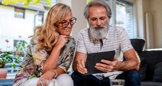 A man and a woman sitting on a couch looking at an ipad that would show information on special enrollment periods for Medicare. Information is not visible. Both are smiling slightly.