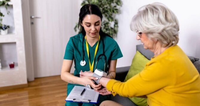 A healthcare worker and a 50+ woman sitting on a couch. The healthcare worker is takin the woman's pulse as part of her Medicare preventive care benefits.